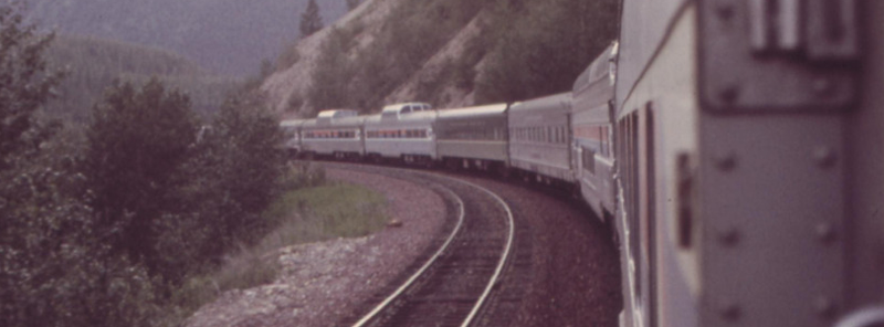 a photograph taken from inside a train, looking along its path, which curves, showing the sides of the cars and the rocky path that the tracks rest on, with trees and mountains surrounding the path