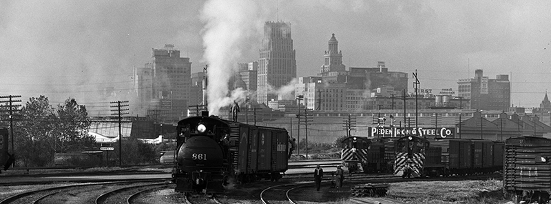 an old black and white photograph of a trainyard where a black train comes towards the viewer, a grand city appearing in the background beyond a row of powerlines and a sign for a steel company