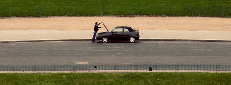 a photograph of a person opening the hood of their car, alone on a road lined on either side by sidewalk and grass