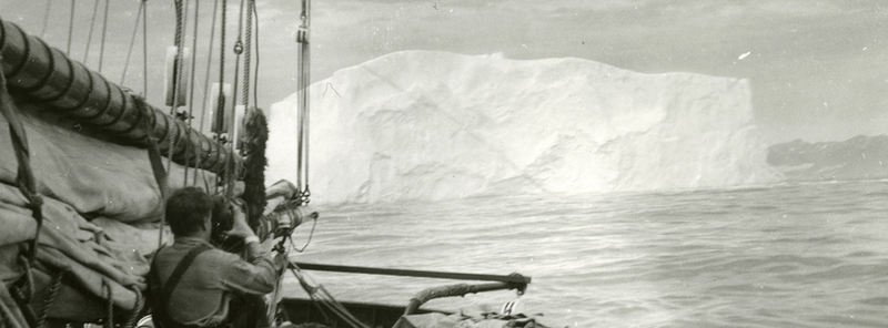 an old black and white photograph of a person using a telescope to peer across the bow of a sailing boat at a large iceberg in the water some 100 or so metres away
