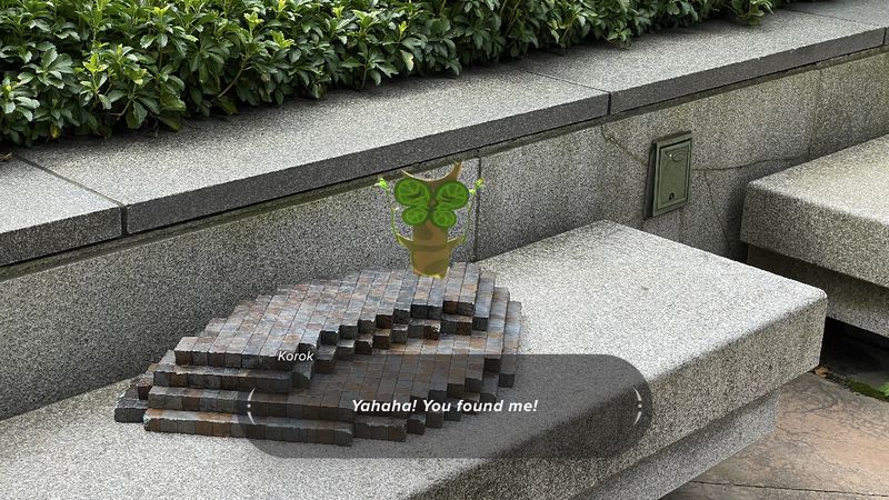 A Korok, resembling a small, living tree with arms, legs, and a smiling face from the video game Zelda: Tears of the Kingdom, stands (superimposed) atop an intricate structure made of small stones, polished into cubes and organised in a tight and symmetrical way. In reference to the video game, where the player can find and replace missing pieces of these structures, and upon doing so, a Korok will appear to congratulate the player, exclaiming, “Yahah! You found me!”, which is displayed in a dialogue box below the Korok.